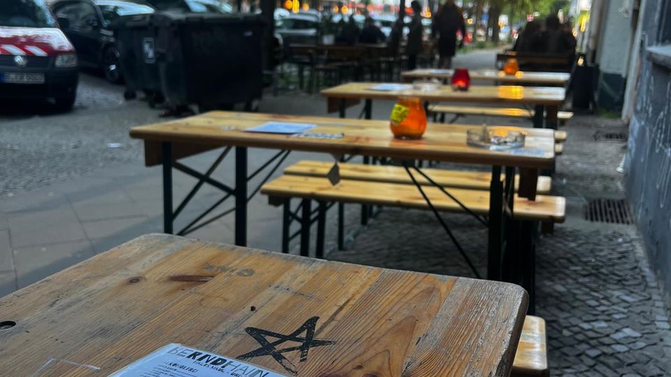 Außenbereich eines Straßencafés mit langen Holztischen und Bänken auf einem Kopfsteinpflaster-Gehweg, auf den Tischen liegen Speisekarten und kleine Kerzen. Entlang der baumbestandenen Straße stehen parkende Autos, und am Gebäude rechts hängt eine ausgefahrene Markise.