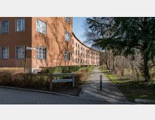 Mehrstöckiges Wohngebäude mit orange-brauner Fassade, das in einem leichten Bogen verläuft. Im Vordergrund verläuft ein gepflasterter Weg durch eine Grünanlage mit Büschen und Bäumen, links ist ein Straßenschild mit der Aufschrift "Am Schäfersee 27-41" sichtbar.