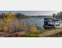Eine Uferpromenade mit Backsteintreppe führt zu einem See, der von herbstlich verfärbten Bäumen gesäumt ist. Im Wasser liegt ein Hausboot, umgeben von weiteren Booten an einem kleinen Hafen.