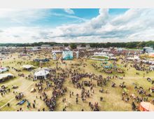 Weitläufiges Festivalgelände mit zahlreichen Zelten, Essensständen, Dekorationen und Menschenansammlungen auf einer Wiese unter einem bewölkten Himmel. Im Hintergrund sind Bühnen, Bäume und weitere Veranstaltungsstrukturen zu sehen.