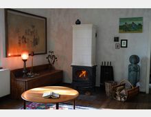 Gemütliches Wohnzimmer mit schlichtem Holzboden, einem weißen Kachelofen mit Feuer, einem runden Holztisch, einer Holzbank und einer beleuchteten Tischlampe. Dekorative Elemente wie ein gemaltes Bild an der Wand, kleine Rahmenbilder, eine Buddha-Statue und ein Kaminholzkorb ergänzen den Raum.