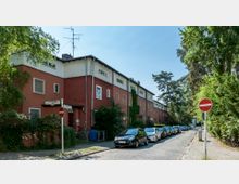 Wohnsiedlung mit rot gestrichenen Reihenhäusern, gesäumt von schmalen Gehwegen und geparkten Autos entlang einer gepflasterten Straße. Umgeben von Bäumen und viel Grün; Straßenschilder und ein "Durchfahrt verboten"-Schild im Vordergrund.