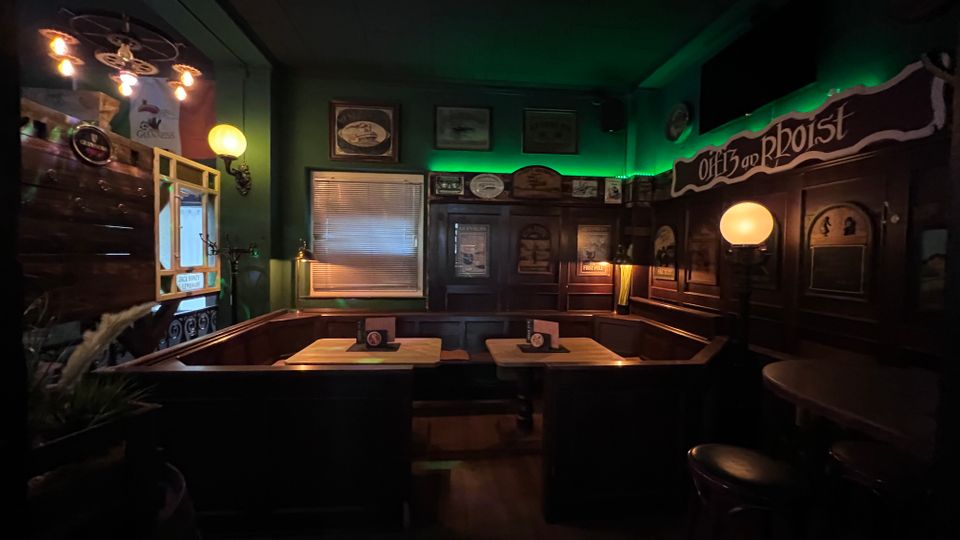 Gemütlicher Pub-Innenraum mit dunkelgrünen Wänden, dunkler Holzvertäfelung und Sitzecken aus Holzbänken um kleine Tische. An den Wänden hängen Schilder und Bilder; warme Kugellampen und ein grüner LED-Lichtstreifen beleuchten den Raum, neben einem Fenster mit Jalousie und einer Tür.