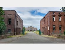 Straßenansicht mit zwei historischen Backsteingebäuden links und rechts, die zu einem zentralen, größeren Gebäude im Hintergrund führen; das Gelände ist von einer Einfahrt mit grünem Tor umgeben, und der Himmel ist teils bewölkt.