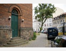 Backsteingebäude mit einer großen grünen Holztür und abgerundeter Fensterverzierung, flankiert von Wandlampen. Im Vordergrund befinden sich ein gepflasterter Gehweg, parkende Autos, ein Baum und eine gelbe Straßenbahn entlang einer ruhigen Straßenszene mit historischen Wohnhäusern im Hintergrund.