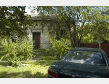 Ein älteres Gebäude mit einer verzierten Holztür, umgeben von Bäumen und Sträuchern. Im Vordergrund steht ein dunkelgrünes Auto auf einer Grasfläche, daneben eine Straßenlaterne.