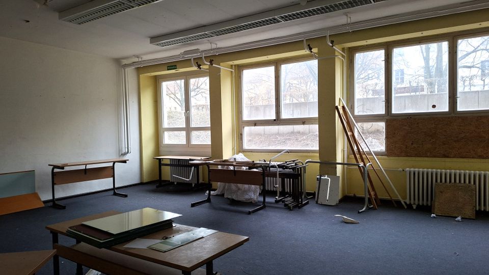 Leerer, teilweise ausgeräumter Klassen- oder Büroraum mit großen Fensterfronten, Heizkörpern darunter und sichtbaren Deckenleuchten. Auf dem Teppichboden stehen einzelne Tische, gestapelte Möbelteile und Holzplatten; draußen sind kahle Bäume durch die Fenster erkennbar.