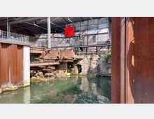 Industriegelände mit offenen Wasserbecken, umgeben von Backsteinmauern und verrosteten Stahlstrukturen. Im Hintergrund sind Geländer, ein modernes Dachgerüst und eine rote Infotafel zu sehen.