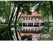 Fachwerkgebäude mit roten Dachziegeln und großen Fenstern, umgeben von Bäumen und einem Wassergraben, in dem sich das Haus spiegelt. Links im Bild sitzen Menschen im Freien auf einer Terrasse, eingerahmt von grüner Vegetation.