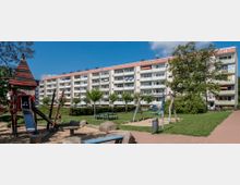 Wohnsiedlung mit einem mehrstöckigen, hell verputzten Wohngebäude im Hintergrund, davor ein gepflegter Spielplatz mit Rutsche, Schaukelgerüst und Sitzbänken sowie Grünflächen und Bäumen. Der Platz ist mit Sand- und Gehwegflächen gestaltet.