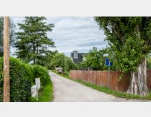 Eine ländliche Straße mit Schotterbelag, umgeben von dichtem Grün, Bäumen und Hecken. Im Hintergrund ist ein modernes Haus mit grauem Dach zu sehen, daneben ein Holzlattenzaun und ein Straßenschild mit der Aufschrift „Schleweg“.