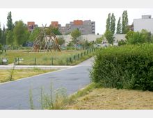 Ein Park mit einer großen Holzspielstruktur in der Mitte, umgeben von Rasenflächen und schmalen Wegen. Im Hintergrund sind mehrstöckige Wohnhäuser sowie einige Bäume und Büsche zu sehen.