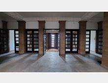 Eingangsbereich eines Gebäudes mit einem symmetrischen Design aus braunen Steinsäulen und verglasten Doppeltüren. Im Hintergrund ist eine moderne Holztür mit kleinen Glasflächen zu sehen, links steht ein Feuerlöscher an der Wand.