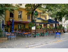Außenbereich eines Restaurants mit dem Namen „Carabao“ vor einem gelben Gebäude mit mehreren Fenstern. Der Bereich umfasst eine Terrasse mit Holzzaun, Tischen und bunten Metallstühlen sowie Sonnenschirmen und Pflanzen als Dekoration.