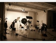 Ein Fotostudio mit mehreren Scheinwerfern, Reflektoren und Stativen, die auf eine weiße Hintergrundwand ausgerichtet sind. Im Studio befinden sich Personen, die mit der Beleuchtung und den Vorbereitungen beschäftigt sind, sowie Kabel und Equipment am Boden.