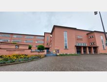 Außenansicht eines Gebäudekomplexes mit orangefarbener Fassade, bestehend aus der "Friedrich-Ebert-Grundschule" und dem "Stadttheater". Links ist ein Schulhof mit Blumenbeeten und rechteckigen Fenstern zu sehen, rechts der Eingangsbereich des Theaters mit hohen Fenstern und Säulenelementen.