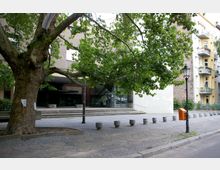 Ein öffentlicher Platz mit gepflastertem Boden und einem großen Laubbaum im Vordergrund. Im Hintergrund ist der Eingang eines modernen Gebäudes mit Glasfassade und Treppenstufen sichtbar; daneben befinden sich Wohnhäuser mit Balkonen.