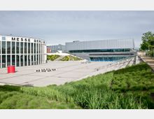 Außenansicht der Messe Berlin mit dem angrenzenden CityCube Berlin, einem modernen rechteckigen Gebäude mit Glasfassade. Im Vordergrund befinden sich eine große gepflasterte Fläche, grüne Rasenflächen und vereinzelt Bänke.