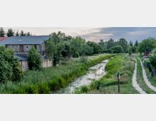 Ländliche Landschaft mit einem schmalen, von dichtem Grün gesäumten Wassergraben, einem Weg mit Grasstreifen und einem alten Backsteingebäude im Hintergrund, umgeben von Bäumen und Sträuchern.