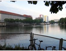 Blick auf einen Fluss mit ruhiger Wasseroberfläche, im Hintergrund ein großes Gebäude mit rotem Dach links und moderne Hochhäuser mit farbigen Akzentfassaden rechts. Im Vordergrund ein Zaun mit angelehnten Fahrrädern, umgeben von Bäumen und Grünflächen.