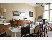 Esszimmer im traditionellen Stil mit gedecktem Tisch, antiken Holzstühlen und Sideboards. Im Raum befinden sich Wandbilder, ein Spiegel, eine Stehlampe und ein Kronleuchter, während Tageslicht durch große Fenster einfällt.