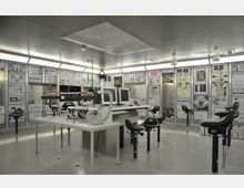 Innenraum eines modernen Labors oder Kontrollraums mit zahlreichen Schalttafeln und technischen Geräten an den Wänden. In der Mitte stehen mehrere Arbeitsplätze mit Monitoren, Tischen und höhenverstellbaren Hockern auf einem metallischen Bodenbelag.