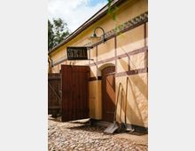 Fachwerkgebäude mit gelblich verputzten Wänden und Holztüren, beschriftet mit einem Schild "Stall". Vor der Gebäudewand stehen landwirtschaftliche Werkzeuge wie Schaufeln und Heugabeln, darunter ein gepflasterter Boden.