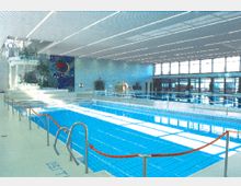 Hallenbad mit einem rechteckigen Schwimmbecken, umgeben von Geländern und roten Absperrseilen. Im Hintergrund sind große Fenster, eine bunt gestaltete Wand mit Mosaik und ein Bereich mit Pflanzen sowie einer Wendeltreppe zu sehen.