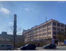 Straßenansicht eines mehrstöckigen, hellbraunen Gebäudes mit zahlreichen Fenstern, daneben ein hoher Industrieschornstein. Im Vordergrund sind parkende Autos, ein Fußgänger, Bäume ohne Laub sowie Büsche zu sehen, bei klarem Himmel.