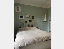 Schlafzimmer mit großem Bett und weißer Bettdecke vor einer salbeigrünen Wand, die mit mehreren gerahmten Bildern dekoriert ist. Neben dem Bett stehen eine schlichte Lampe und Bücher, rechts ist ein kleines tiefes Fenster in der Wand zu sehen.