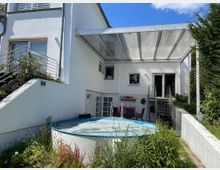 Einfamilienhaus mit weißer Fassade, einer überdachten Terrasse und einer Außentreppe, die zu einem Garten mit einem kleinen, runden Aufstellpool und umliegender Vegetation führt. Im Hintergrund stehen Gartenmöbel unter der Überdachung.