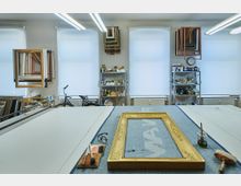 Ein Atelierraum mit großen Fenstern und geschlossenen Rollos, mehreren Regalen voller Materialien und Werkzeugen sowie aufgehängten Holzrahmen. Im Vordergrund liegt ein vergoldeter Bilderrahmen auf einem Arbeitstisch mit Pinseln und anderen Restaurierungsutensilien.