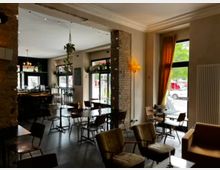 Innenraum eines gemütlichen Cafés mit Holzmöbeln, sichtbaren Backsteinwänden und großen Fenstern, die Tageslicht hereinlassen. Die Einrichtung umfasst Hängelampen, Pflanzen und eine Bar im Hintergrund mit Barhockern.