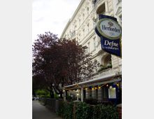 Mehrstöckiges historisches Wohngebäude mit verzierten Fassaden und Balkonen; im Vordergrund ein Restaurant mit überdachter Terrasse, dekoriert mit Lichterketten. Ein Baum mit dunkelrotem Laub und ein Werbeschild des Restaurants ragen in die Szene.