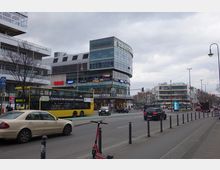 Straßenansicht einer belebten Kreuzung mit mehrspurigen Fahrbahnen, einem gelben Doppeldeckerbus und mehreren Autos. Im Hintergrund ein modernes Einkaufszentrum mit Glasfassade, leuchtenden Schildern von Geschäften wie Primark, REWE und dm sowie umliegenden Bürogebäuden.