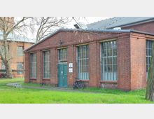 Backsteingebäude mit großen Sprossenfenstern und einem grünen Doppeltor, beschriftet als "Gebäude 23". Im Vordergrund eine kleine Rasenfläche, im Hintergrund Bäume und ein weiteres Gebäude aus rotem Backstein.