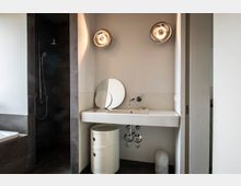 Modernes Badezimmer mit weißem Waschbecken, kugelförmigen Wandlampen, einem runden Spiegel und dunklen Fliesen. Links befindet sich eine Duschkabine mit Regenduschkopf und Glastrennwand, daneben eine freistehende kleine Kommode und ein Mülleimer unter dem Waschbecken.