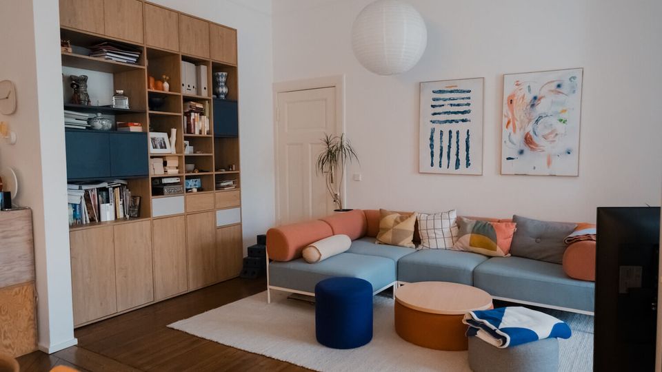 Bright, modern living room with a built-in wooden shelving wall filled with books and decor, a pale blue sectional sofa with colorful cushions, and a low round coffee table on a light area rug. White walls feature two abstract framed artworks, with a paper globe pendant light overhead and a small plant near a closed door.