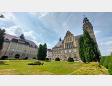 Historisches Gebäudeensemble mit zwei großen Gebäuden: Links ein sandfarbenes Gebäude mit dunklem Dach, rechts eine höhere, steinerne Fassade mit Turm und Uhr. Dazwischen befindet sich eine gepflegte Rasenfläche mit Blumenbeeten und hohen Bäumen. Der Himmel ist leicht bewölkt.