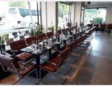 Ein modernes Restaurant mit großen Fensterfronten, durch die eine Straße mit parkenden Fahrzeugen sichtbar ist. Im Innenraum stehen lange, gedeckte Tische mit braunen, gepolsterten Stühlen auf einem Terrazzo-Boden; auf den Tischen befinden sich Gläser, Besteck, und Blumen.