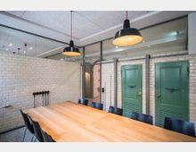 Moderner Besprechungsraum mit hellem Holztisch und schwarzen Stühlen, umgeben von weißen Fliesenwänden. Im Hintergrund befinden sich zwei grüne Türen und eine wandhohe Glasfläche mit Blick in einen benachbarten Bereich; von der Decke hängen schwarze Pendelleuchten.