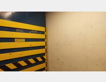 Innenansicht eines Lastenaufzugs mit blauer Wand und gelben Querlatten, die durch schwarze Streifen markiert sind. Die gegenüberliegende Wand ist hell und mit Kritzeleien versehen; ein Schild zeigt eine maximale Kapazität von 20 Personen oder 1500 kg an.