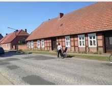 Straßenszene in einem Dorf mit Fachwerkhäusern aus Ziegelstein und roten Ziegeldächern, im Vordergrund eine Person, die ein Fahrrad schiebt. Die Straße ist mit Kopfsteinpflaster bedeckt und von schmalen Gehwegen und Grünstreifen gesäumt.