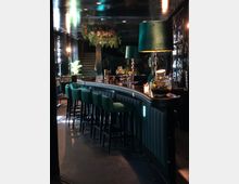 Eine stilvoll eingerichtete Bar mit dunklem Interieur, grünen Polsterhockern und einem geschwungenen Tresen. Im Hintergrund sind Regale mit Flaschen, eine Treppe und dekorative Pflanzen, einschließlich hängender Blumenarrangements, zu sehen.