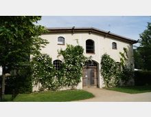 Zweistöckiges Haus mit cremefarbener Fassade und Holztor, bewachsen mit rankenden weißen Blumenrosen. Umgeben von gepflegtem Rasen, Bäumen und Büschen, unter klarem Himmel.