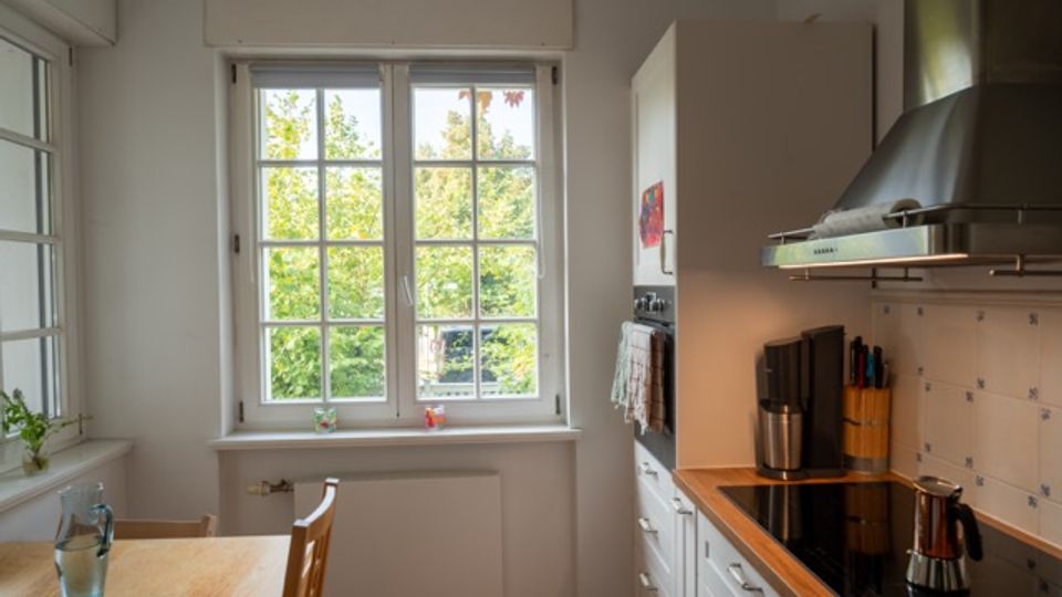 Helle, schmale Küche mit weißer Einbauzeile und Holzarbeitsplatte, Cerankochfeld sowie Edelstahl-Dunstabzugshaube an einer gefliesten Wand. Ein großes Sprossenfenster mit Blick ins Grüne und ein kleiner Esstisch am Fenster prägen den Raum.