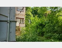 Außenansicht eines Gebäudes mit hellem Putz und braunen Fensterrahmen. Eine schmale Treppe führt durch dichten Bewuchs aus Sträuchern und Bäumen hinauf zur Eingangstür, die von großen Fenstern umgeben ist.