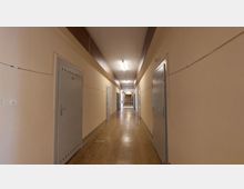 Langer, schlichter Flur in einem Gebäude mit beigen Wänden und einem braunen Linoleumboden. Links und rechts sind graue Türen mit Nummerierungen angebracht, die Decke ist mit länglichen Leuchtstofflampen beleuchtet.