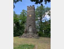 Steinerner Turm aus dunklem Bruchstein mit zinnenartigem Abschluss, umgeben von Bäumen und Grünfläche. Der Turm steht auf einem Sockel aus großen Felsblöcken und zeigt ein eingelassenes, rundes Relief in der oberen Hälfte.