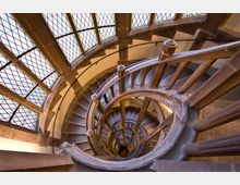 Spiralförmiges Treppenhaus aus Stein in einem historischen Gebäude, mit einem Geländer aus Rundsäulen und großen Fenstern mit rautenförmigen Gittermustern, die Tageslicht hereinlassen. Blick von oben in die Tiefe des Treppenschachts.
